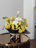 Yellow roses and white blooms with greenery in dark wrapping tied with a champagne-coloured ribbon.