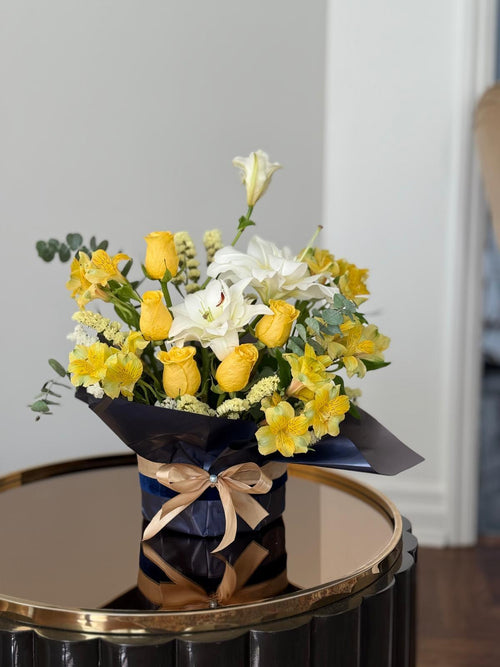 Yellow roses and white blooms with greenery in dark wrapping tied with a champagne-coloured ribbon.