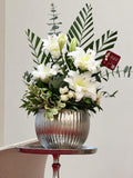 White lilies and small white blooms with eucalyptus and tall palm fronds in a ribbed silver-toned vessel.